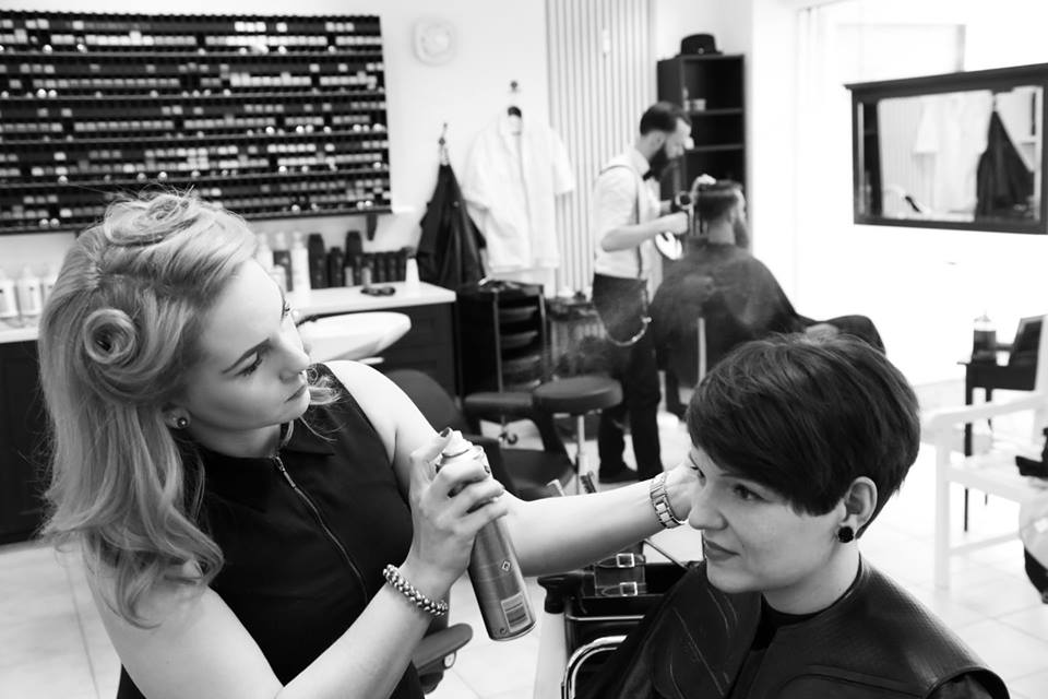 Maria, die Friseurmeisterin und Inhaberin des Salons, steht im Salon und frisiert einer Frau mit kurzen, dunklen Haaren ihre Bobfrisur. Sie benutzt dabei Haarspray. Im Hintergrund sieht man den Salon und einen Friseur, der ebenfalls einem Kunden die Haare macht. Das Bild ist in schwarz-weiß.