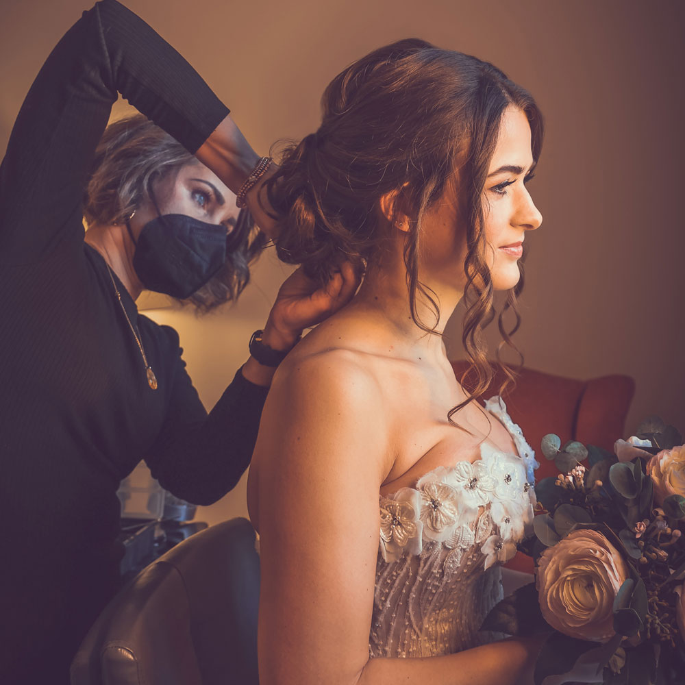 Maria, Friseurmeisterin und Inhaberin des Salons, ist schwarz gekleidet und steckt einer Braut im Brautkleid mit Blumenstrauß und braunen Haaren, die Haare gerade zu einer lockeren Hochsteckfrisur.