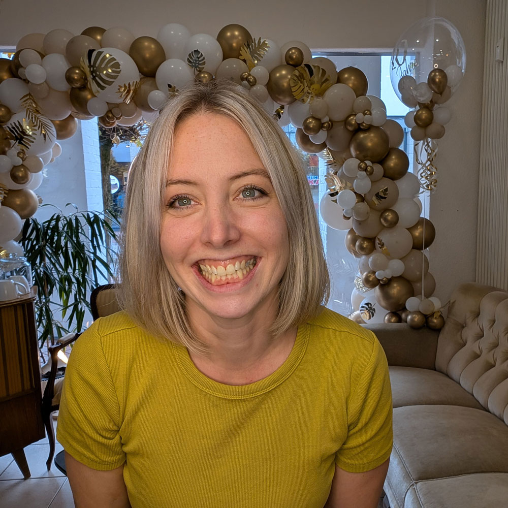 Auf dem Foto sieht man Lisa, Friseurmeisterin und eine Frau Mitte 30. Sie hat mittellange blonde Haare und trägt ein gelbes Shirt. Sie hat ein strahlendes Lächeln und zeigt Zähne.