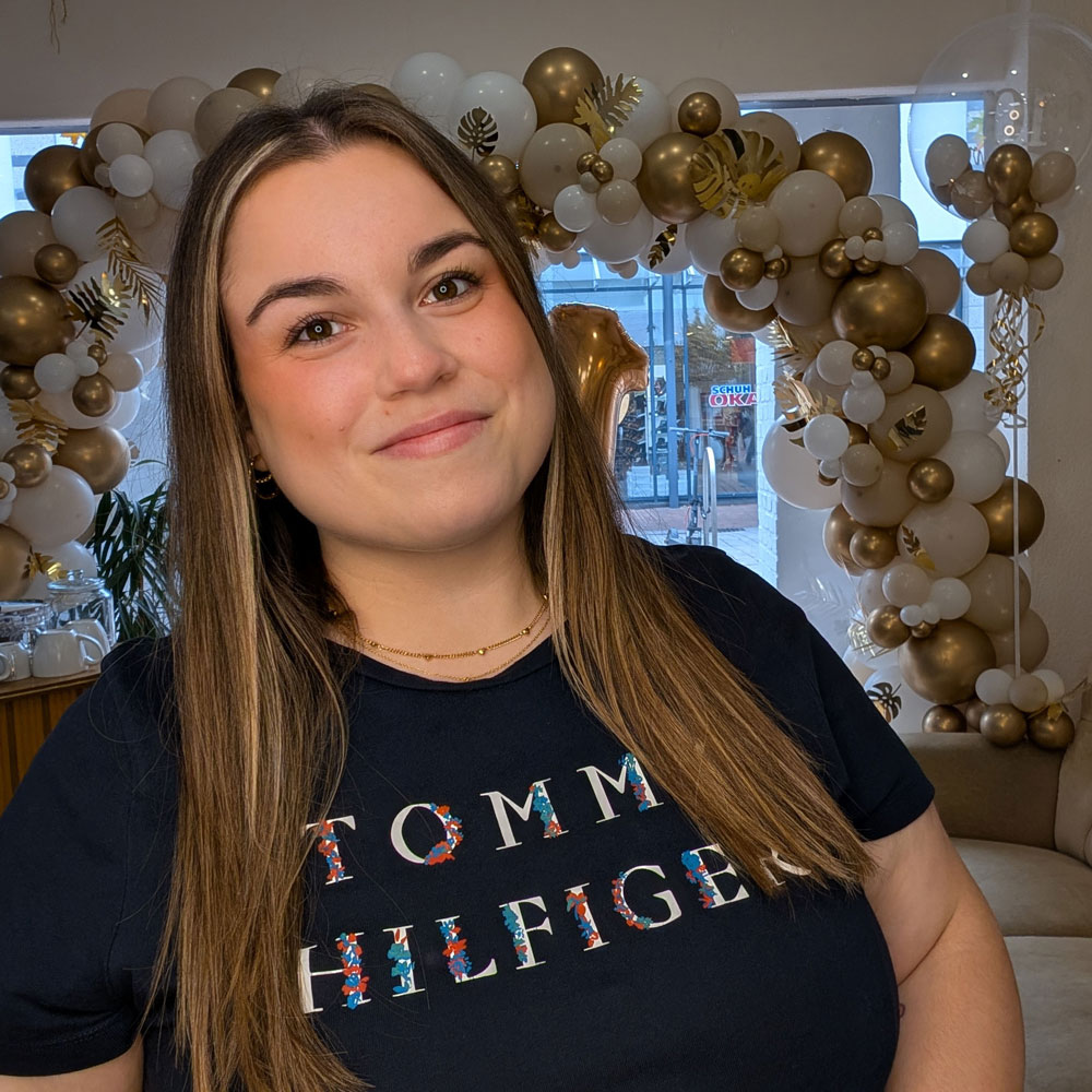 Auf dem Foto ist Marcia, eine Frau mit 20, zu sehen. Sie trägt ein blaues Shirt. Sie hat offene Haare, einen ebenmäßigen Teint und zeigt ein breites Lächeln. Sie hat braune Augen. Im Friseursalon die Stube ist sie Friseurin.