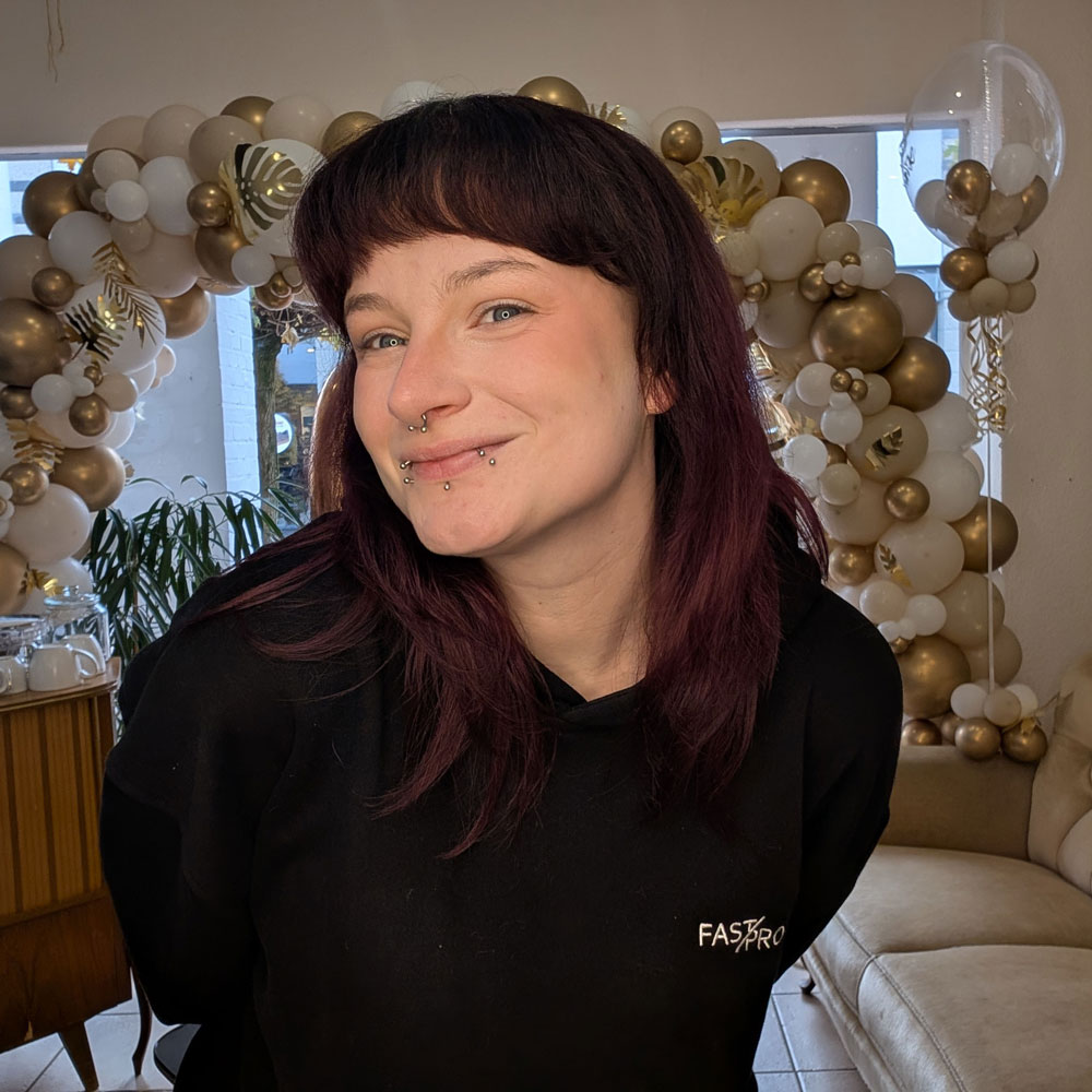 Auf dem Foto sieht man Monique, eine junge Frau Mitte 20. Sie trägt einen schwarze Hoodie. Sie hat dunkelrote Haare, einen Pony. Sie hat zudem Sommersprossen und lächelt verlegen.