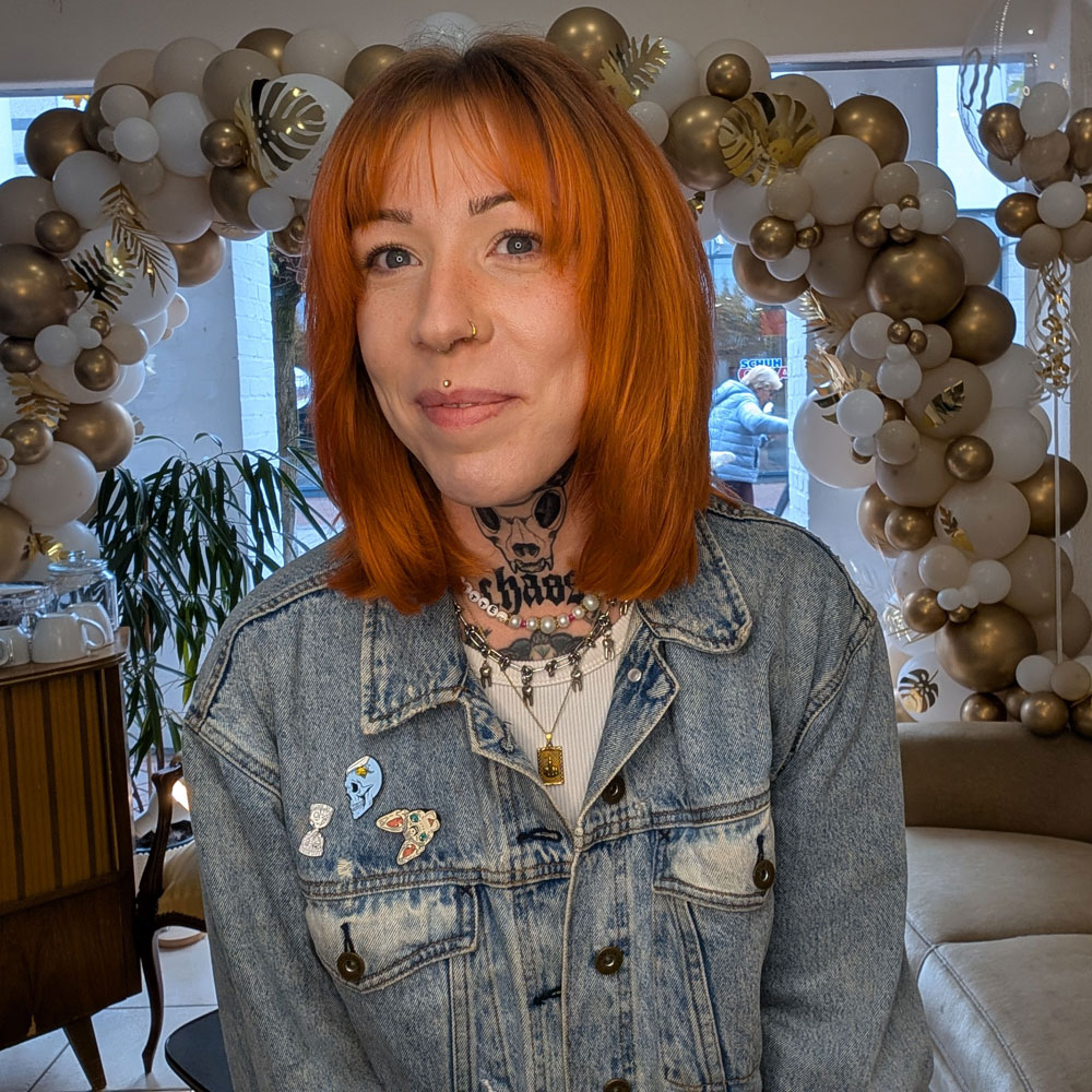 Auf dem Foto sitzt eine Frau Mitte 30 auf einer beigen Couch. Sie heißt Liz und ist Friseurin in der Stube in Emsdetten. Sie trägt eine Jeansjacke. Sie hat einen Mittelscheitel, hat kupferrote Haare, Curtainbangs. Sie lächelt und sie hat ein Nasenpiercing und eins über der Lippe.