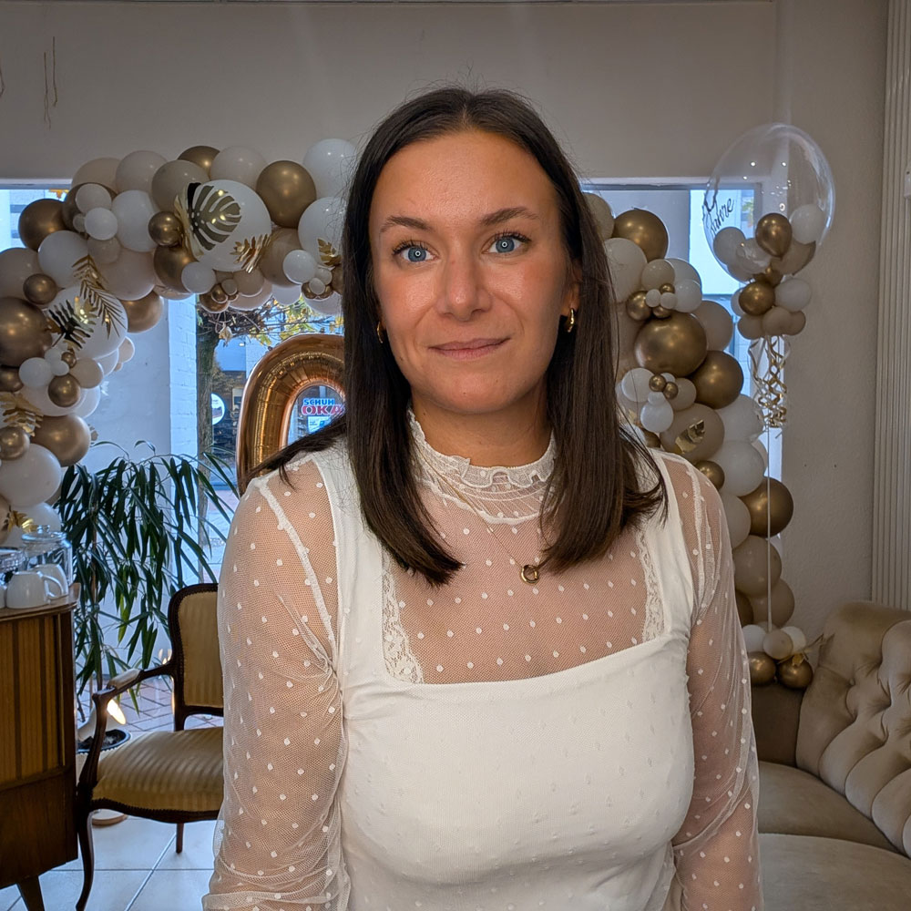 Auf dem Foto sitzt eine Frau, Mitte 30, Friseurin in der Stube in Emsdetten auf einer beigen Couch. Sie weißes Spitzenoberteil. Sie hat offene, braune und schulterlange Haare. Sie lächelt freundlich.