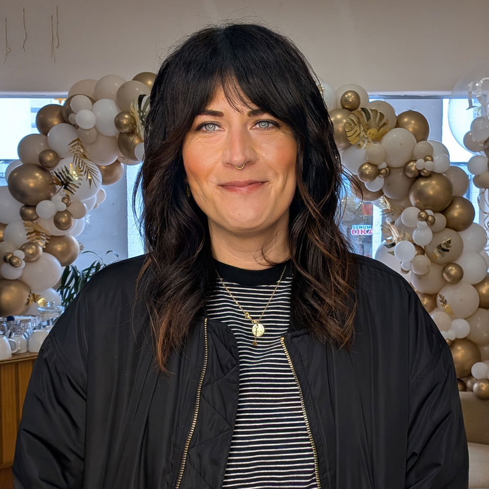 Auf dem Bild sieht man Laura, sie ist Friseurin im Friseursalon die Stube. Sie sitzt auf einer Couch und trägt einen Braunen Pullover, eine schwarze Jacke und Ringelshirt, wie auch eine goldene Kette. Sie lächelt und hat strahlend Blaue Augen und dunkle, offene, Schulterlange Haare.