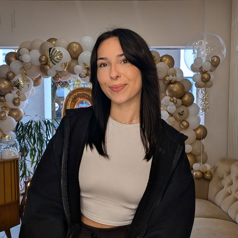 Auf dem Portrait ist die Friseur-Azubine Marita zu sehen. Sie sitzt auf der beigen Couch im Salon, lächelt in die Camera und trägt ein graues Shirt und einen schwarze Pullover. Ihre Haare sind dunkelbraun und offen und sie lächelt in die Kamera.