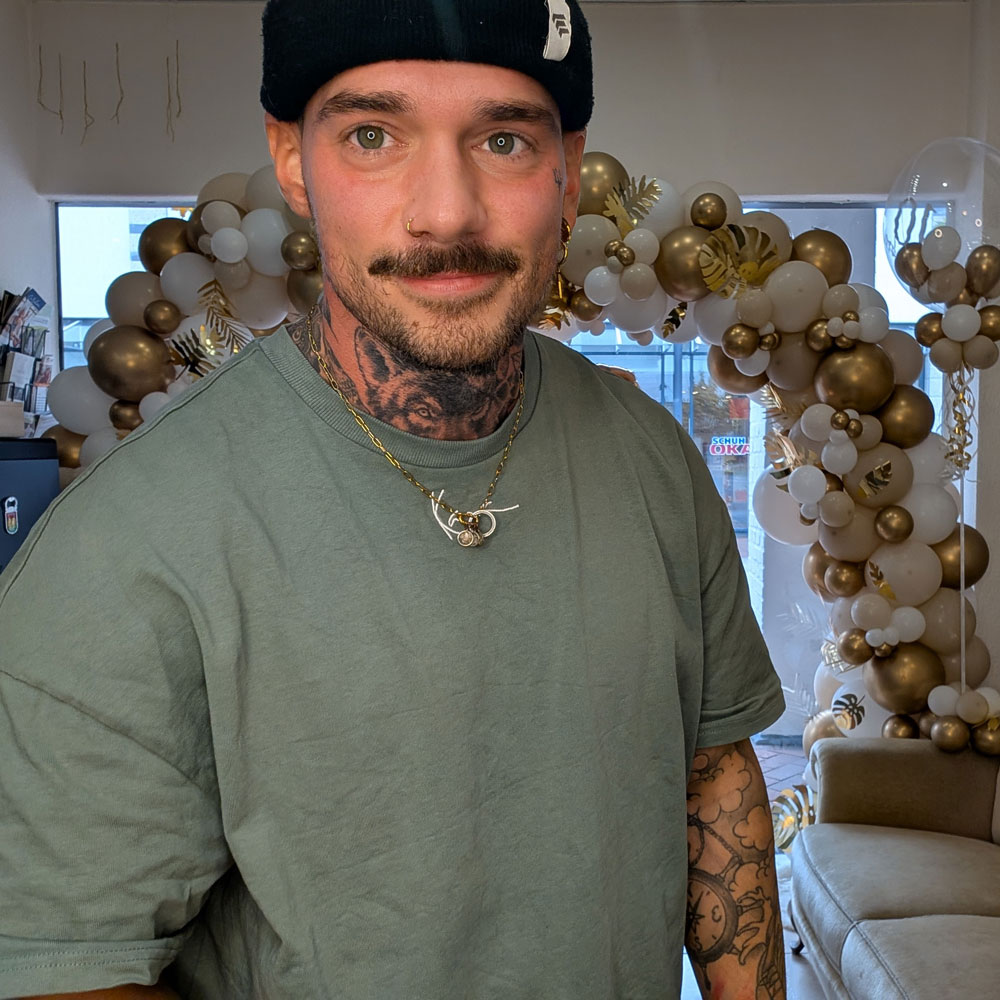 Jan ist ein nett aussehender, stark tattowierter Mann, mit Mintfarbenem Shirt, der eine Mütze trägt. Er hat ein Nasenpiercing, einen Bart und lächelt die Kamera
